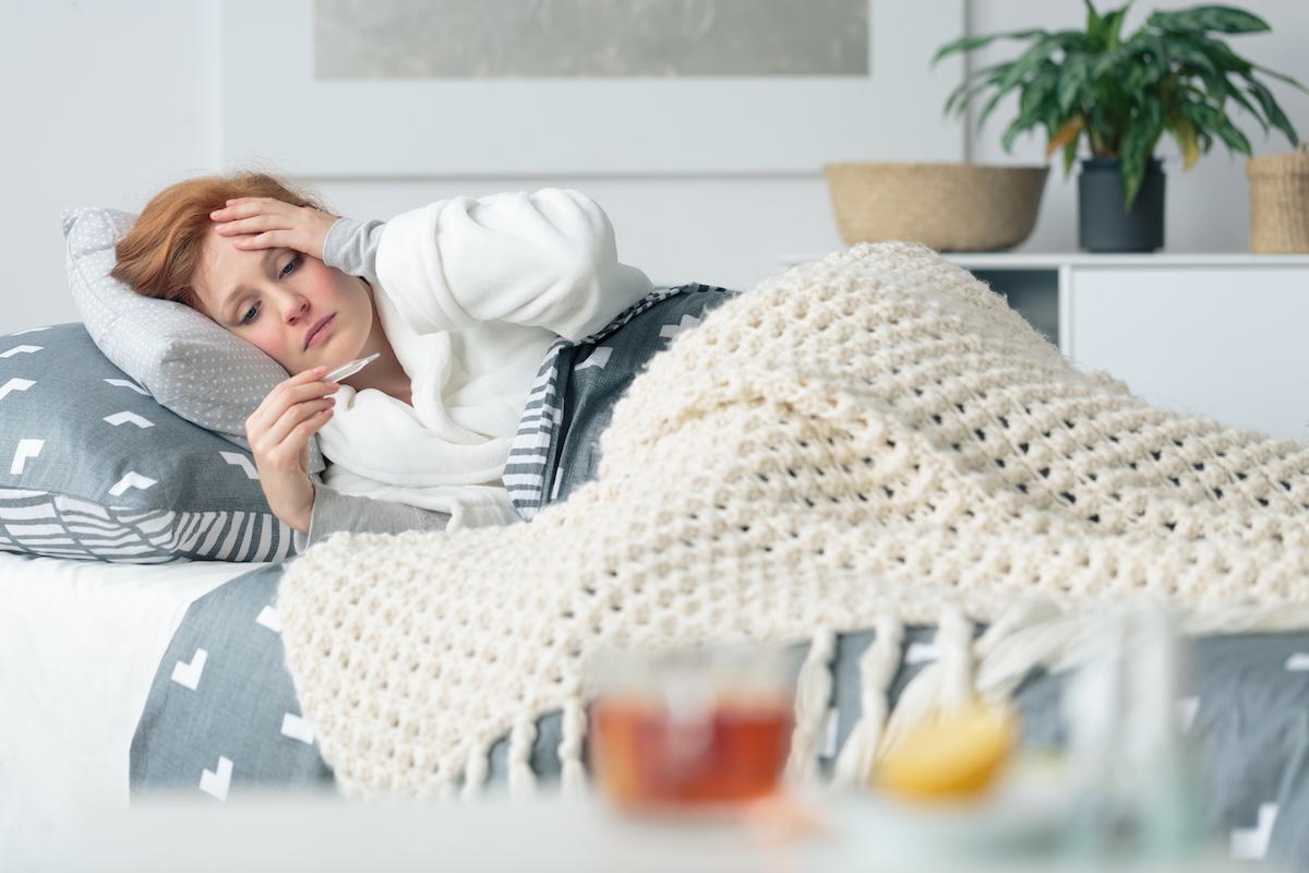 Ill woman lying in bed, looking at a thermometer while recovering from illness