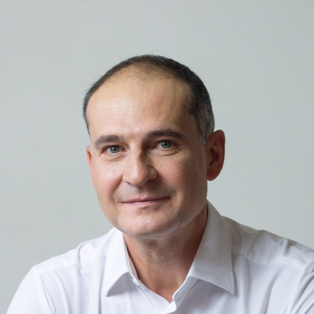 Headshot of Slav Marinov wearing a white button up shirt, against a grey background