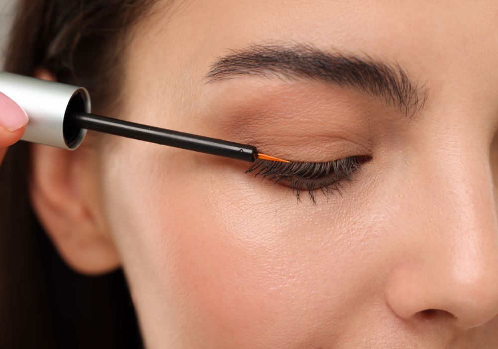 A close up photo of a white woman applying lash serum to her eyelashes