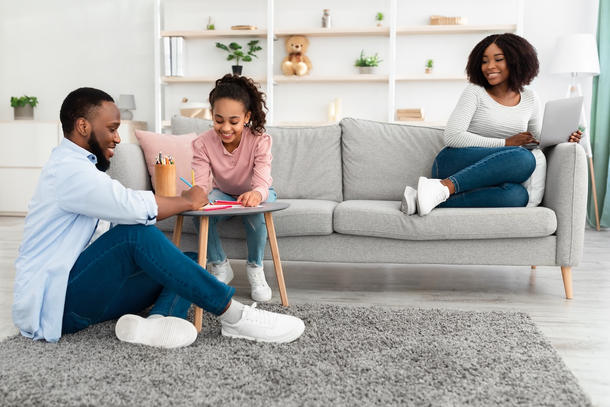 Smiling woman working online on a laptop from home while father plays with daughter, showing work-life balance for beauty business owners