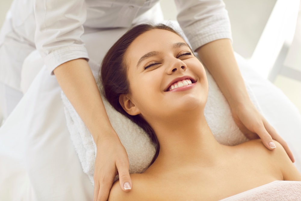 Client lying on a treatment bed, smiling after a facial, with therapist’s hands on their shoulders showing care and relaxation