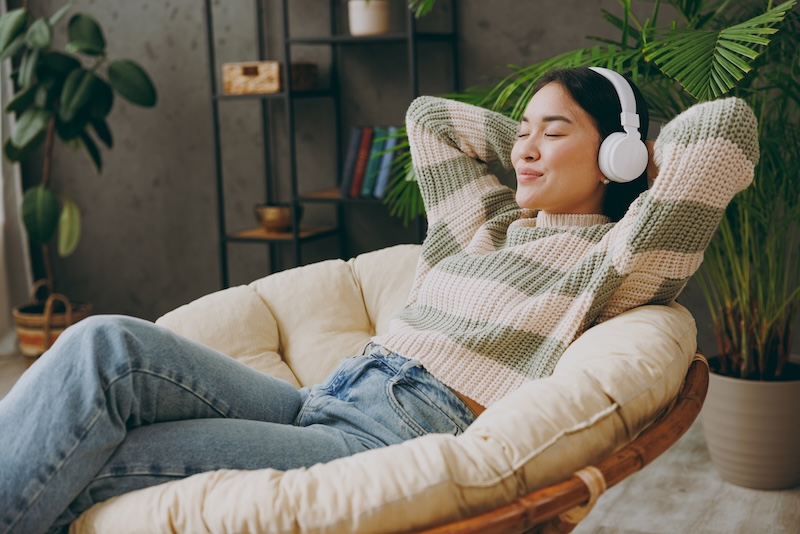 Young woman wearing headphones, sitting on an armchair with eyes closed, relaxing and listening to a podcast