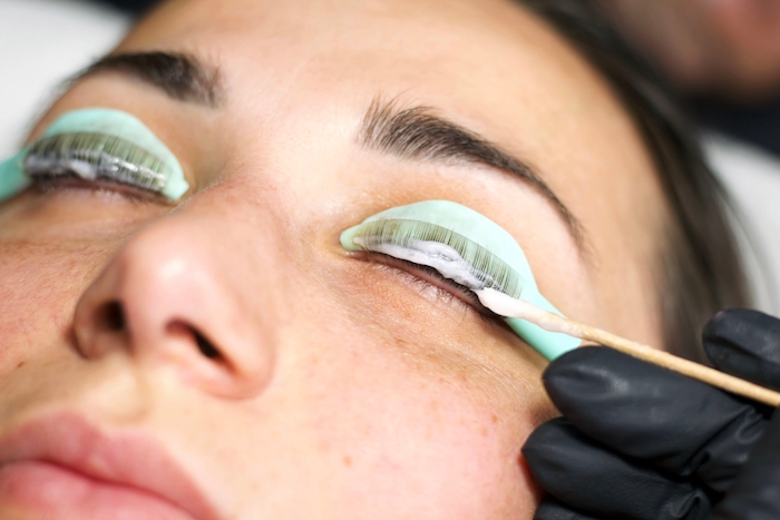 Woman lying down with hey eyes closed while a beauty professional applies a perming solution for a lash lift