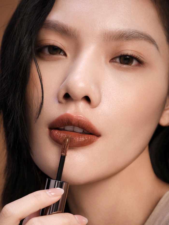 Model with minimal skin and eye makeup, applying glossy brown lipstick