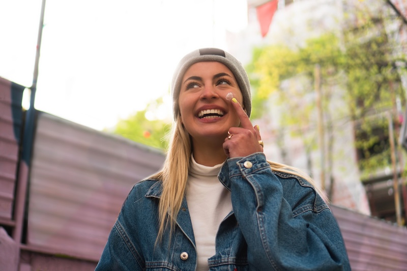 Smiling young woman applying face cream outdoors in an urban setting, representing antioxidant skincare protection against pollution and environmental stress