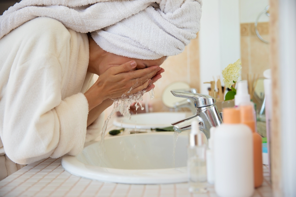 Woman in a white robe and her hair in a towel, washing her face at a basin