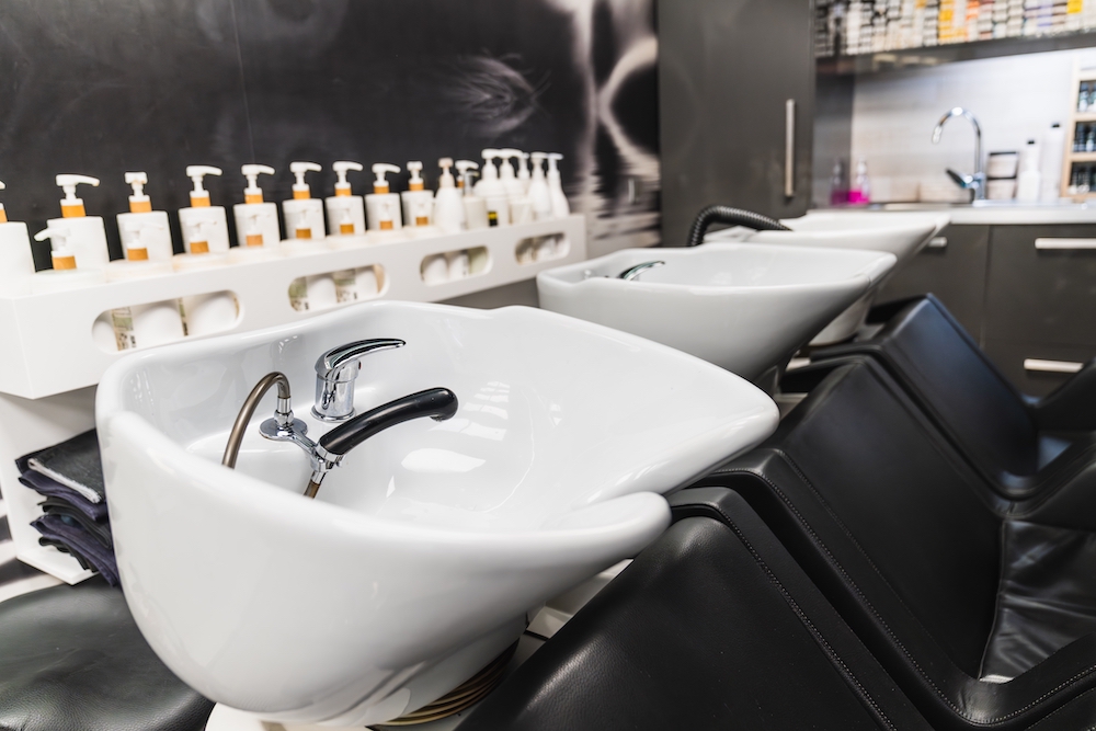 Empty backwash basins in hair salons, with black leather chairs and products lined up on counter at the back