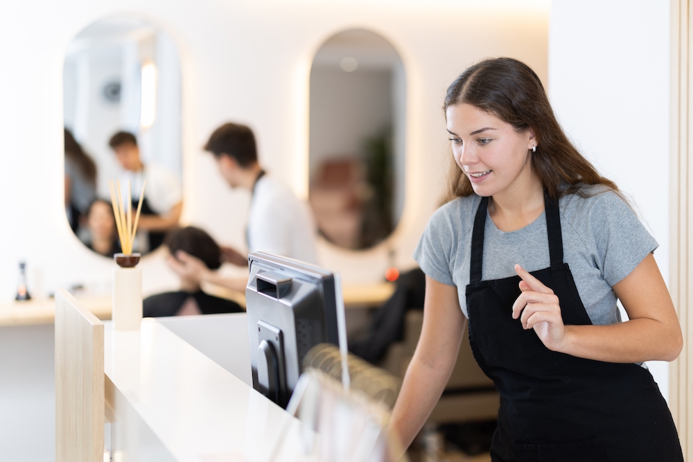 Young hair salon business owner standing at reception desk looking at computer