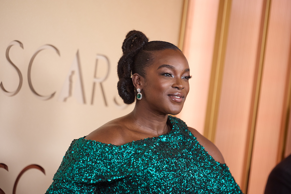 Actress Wunmi Mosaku wearing a shimmering emerald green off-the-shoulder gown and matching green earrings smiles while posing on a red carpet in front of an Oscars backdrop. Her hair is styled in an elegant updo.