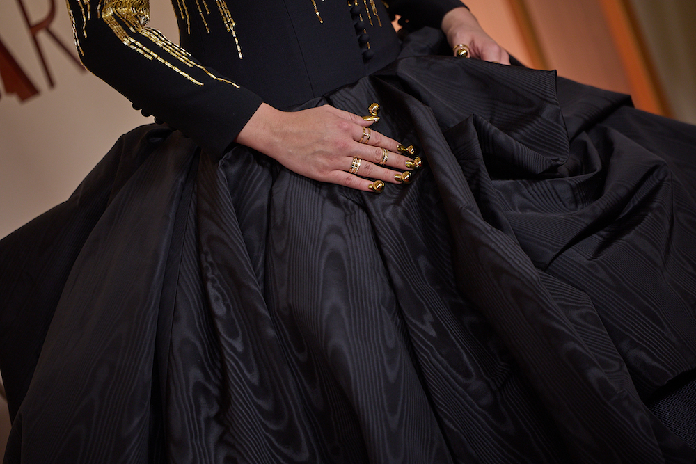 Close-up of Audrey Nuna&rsquo;s hand resting on a voluminous black gown, showing gold metallic manicure and multiple gold rings, with gold detailing visible on the bodice above.