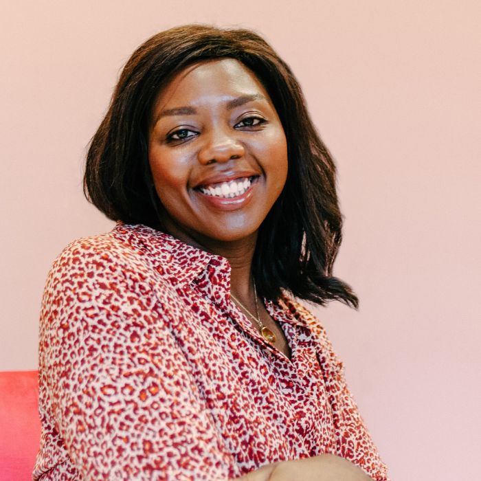Headshot of Dija Ayodele in a red and white animal print blouse, against a pink background