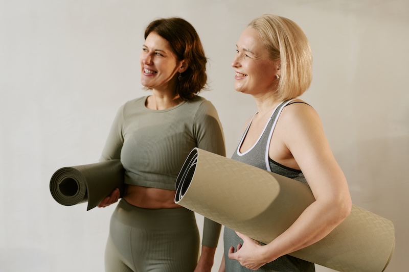 Two menopausal women holding exercise mats, preparing for a Pilates or yoga session to support strength, mobility, and wellness