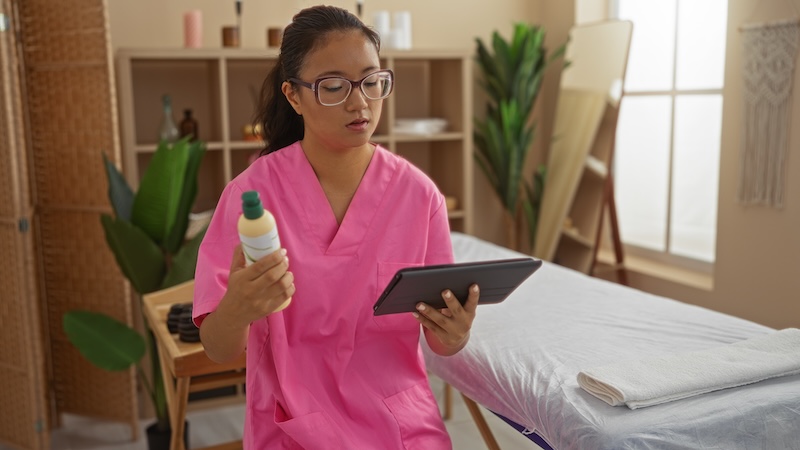 Beauty salon owner reviewing stock levels and comparing a product with a tablet, illustrating cost tracking and operational pressures linked to rising material costs and inflation in the UK beauty industry.