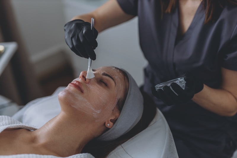 Skin therapist applying an acid mask to a client’s face using a brush during a professional facial treatment