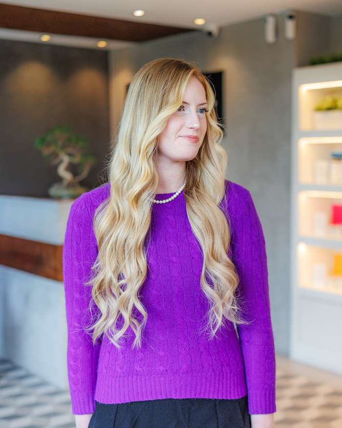 Client at The Hair Movement Salon, standing in reception wearing a purple jumper, with long blonde hair curled and fitted with BHBD hair extensions
