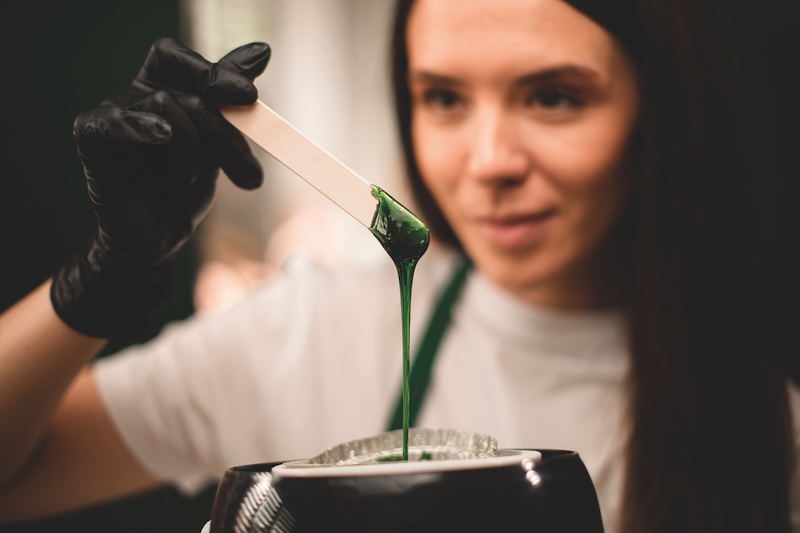 A waxing specialist checking wax in a heater pot during salon treatment preparation, focusing on temperature control and professional technique