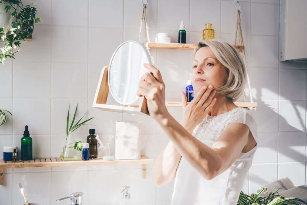 woman checking her skin in a mirror while applying skincare products at home.