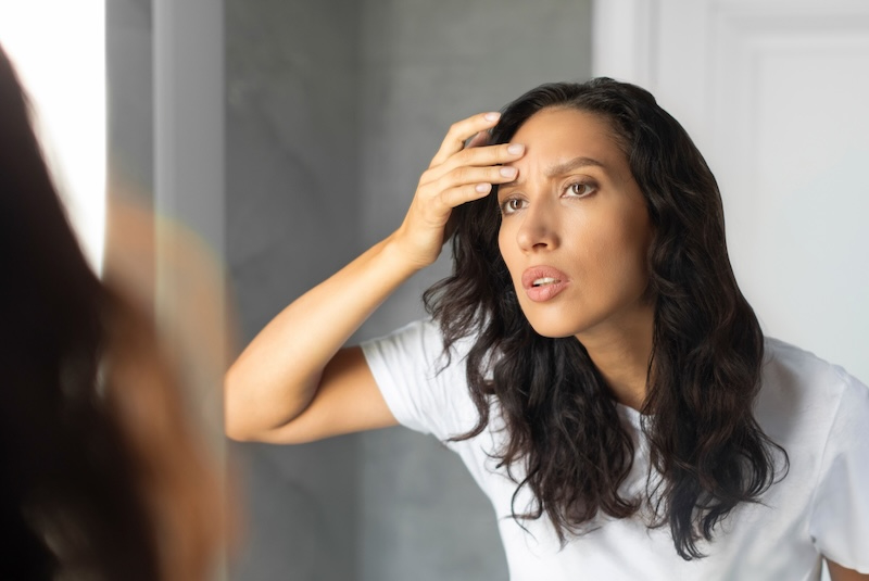 Woman examining fine lines on her forehead while looking in a mirror