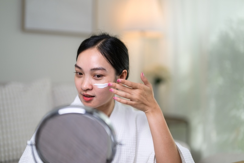 Woman applying facial moisturiser to her cheek while looking in a mirror.