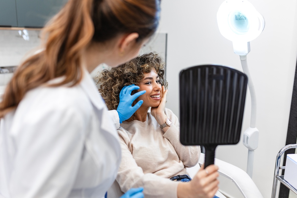 Practitioner assessing a smiling patient’s facial features during an aesthetic consultation using a handheld mirror.