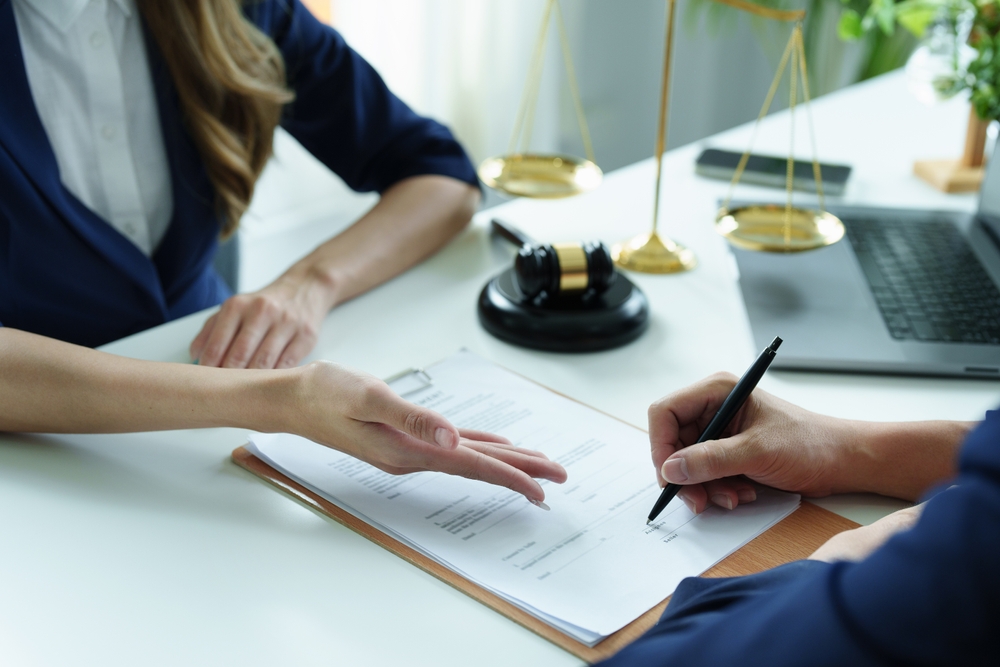 A person signing a legal document at a desk, with a judge’s gavel and scales of justice in the background.