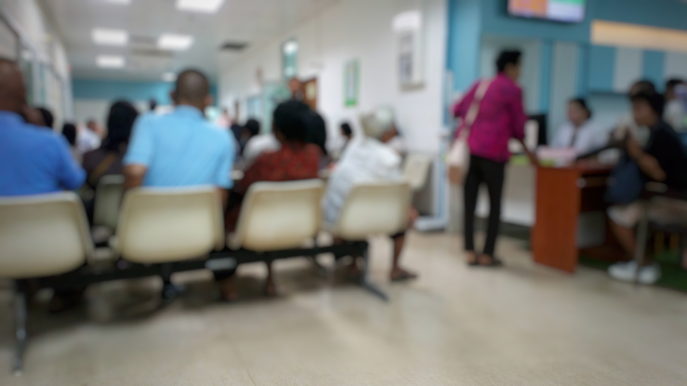 A blurred hospital or healthcare waiting room with patients seated in rows and people speaking with staff at a reception desk.