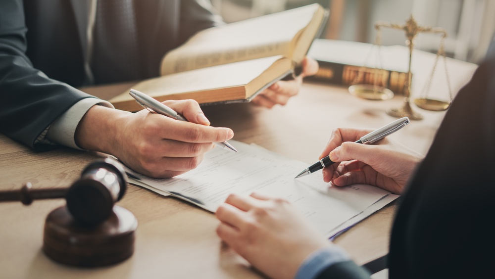 Two people signing legal documents at a desk, with law books, a judge’s gavel and scales of justice in the background.