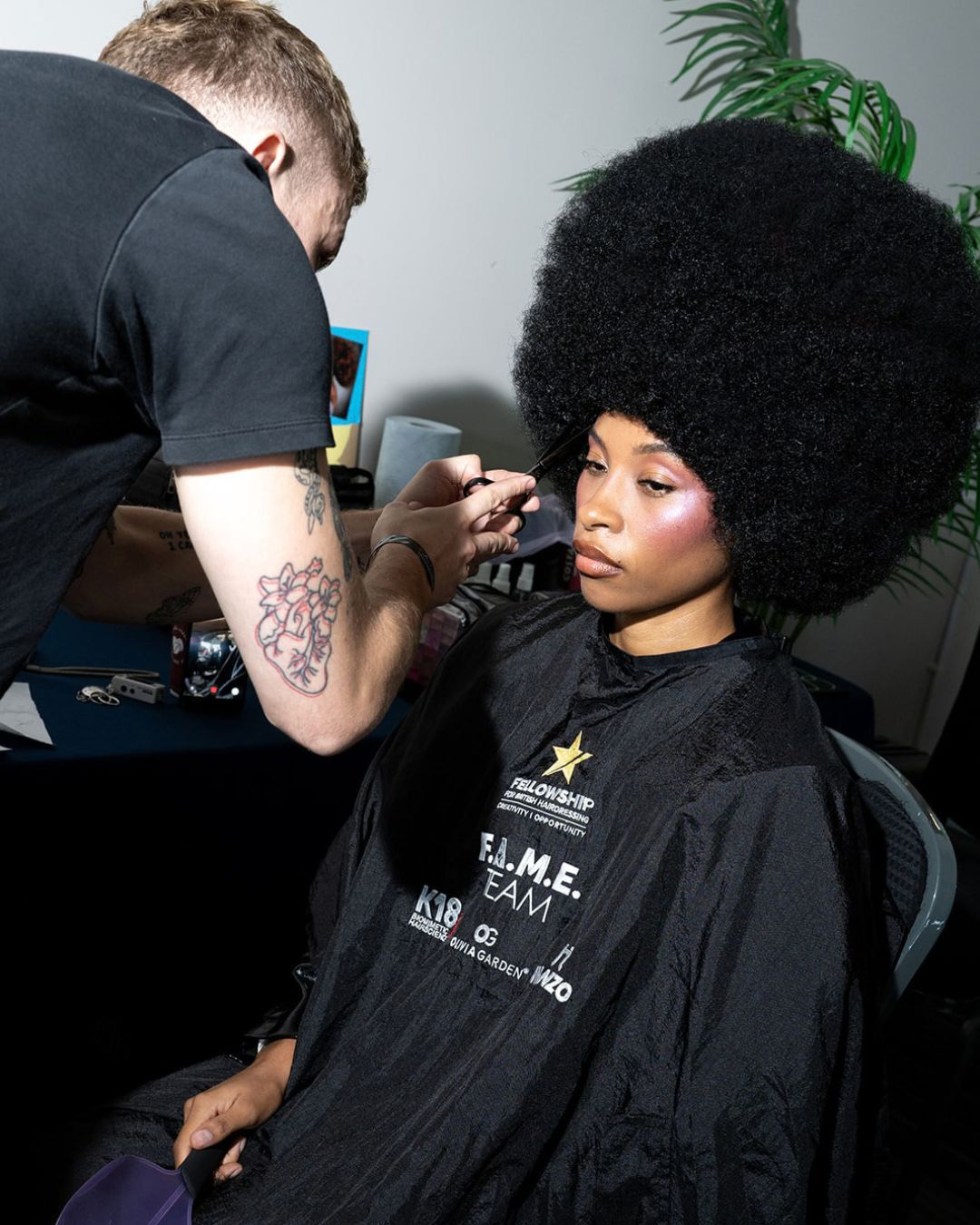 A hairdresser carefully styling a model's voluminous afro hairstyle backstage at the Fellowship F.A.M.E. Team Finals during Salon International 2026.