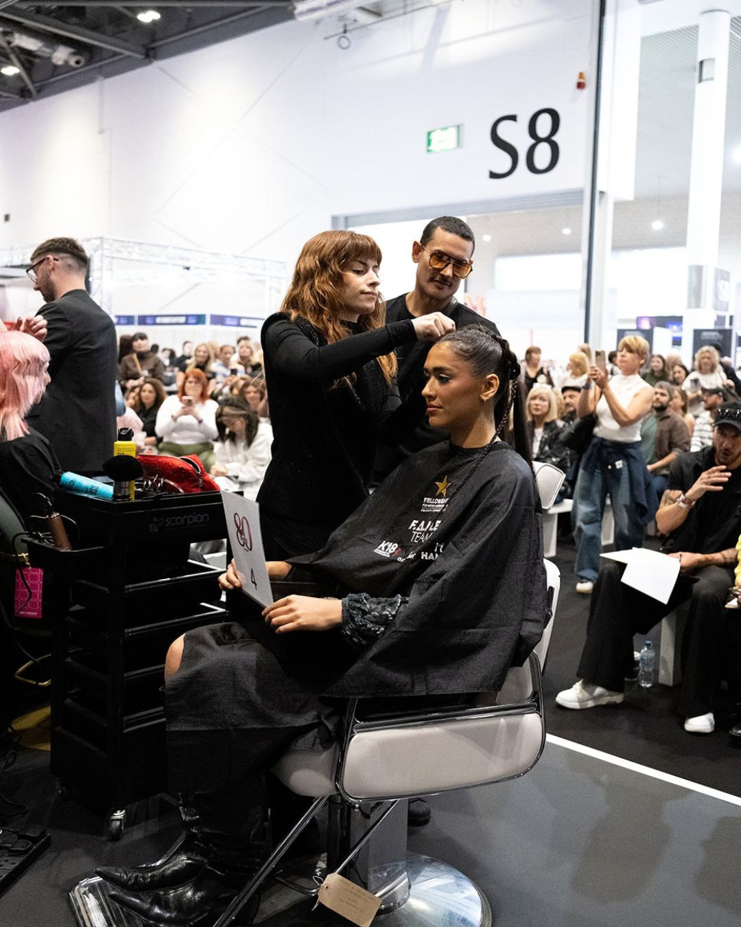 A F.A.M.E. Team finalist styling a model's intricate braided updo live on the HJ Trends Stage at Salon International 2026, watched by judges and an audience.